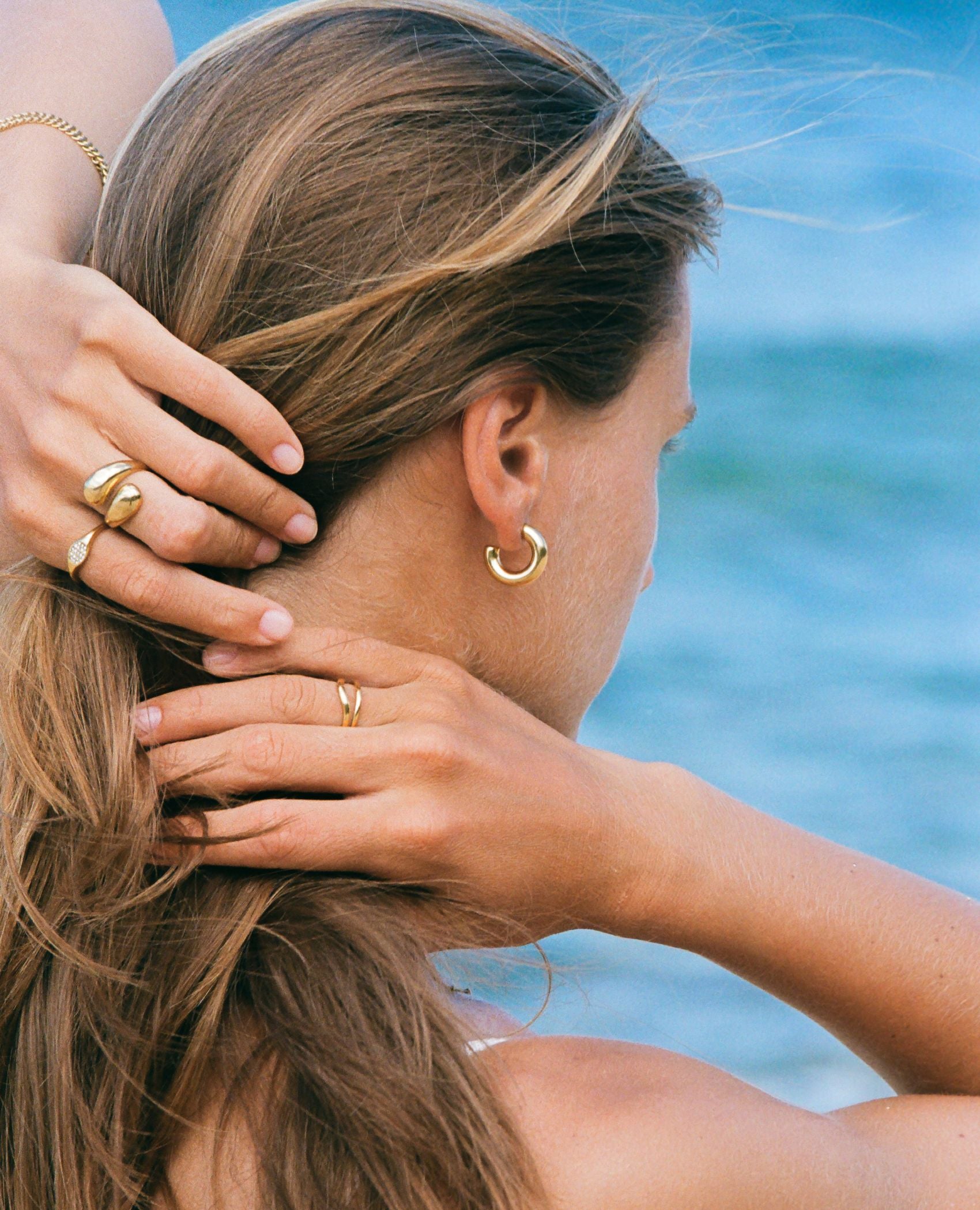 Person at the beach with gold jewelry on their hand and ear.