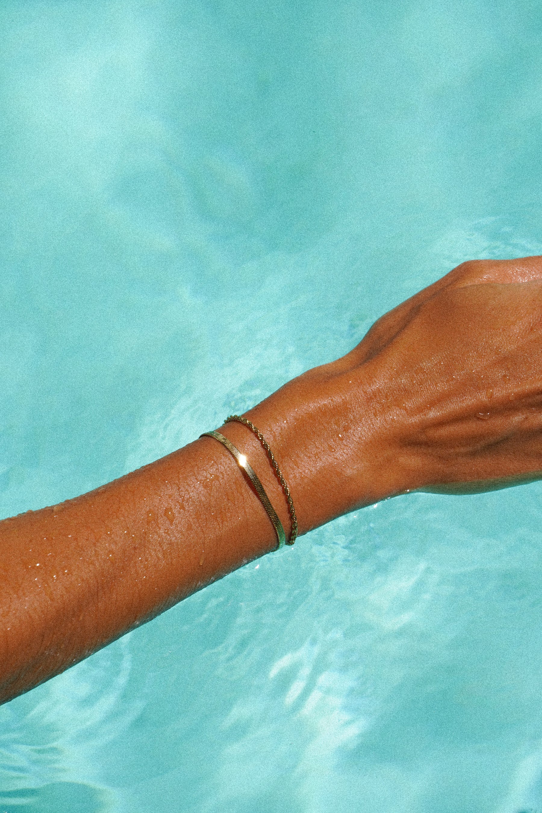 A tanned arm with two gold bracelets in a turquoise pool.